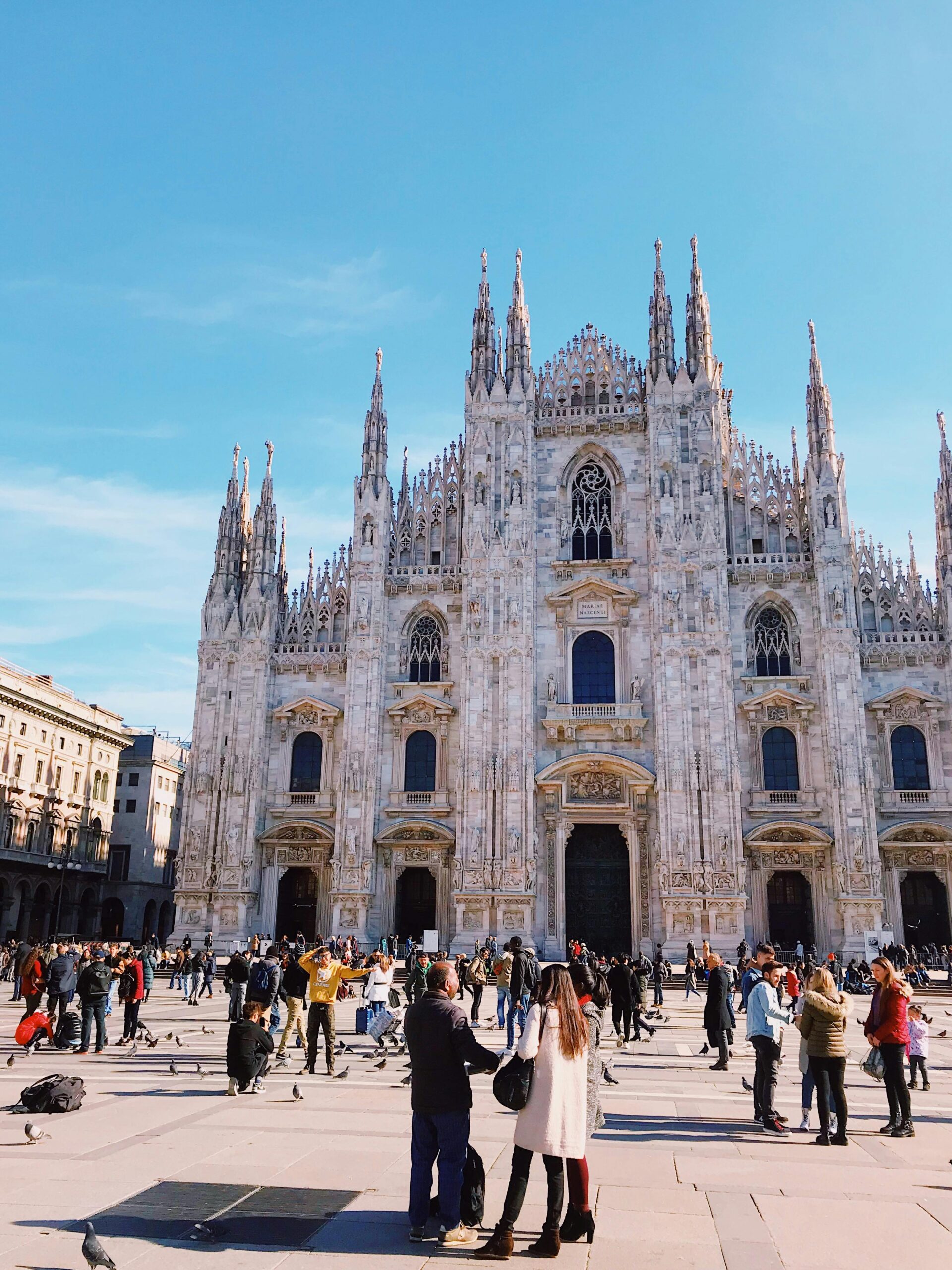 The stunning Milan Cathedral under bright skies, surrounded by a lively crowd in the bustling Piazza del Duomo.