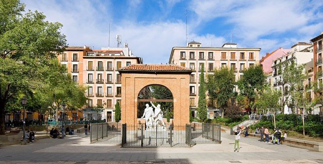 A panoramic view of a city square featuring a fountain and statue, with a skyline of buildings and streets in the background.