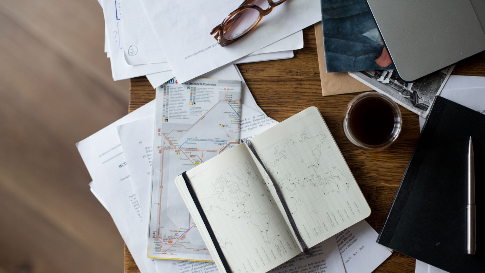 Flat lay of a trip planning workspace with a laptop, folded maps, notebook and coffee on a wooden desk