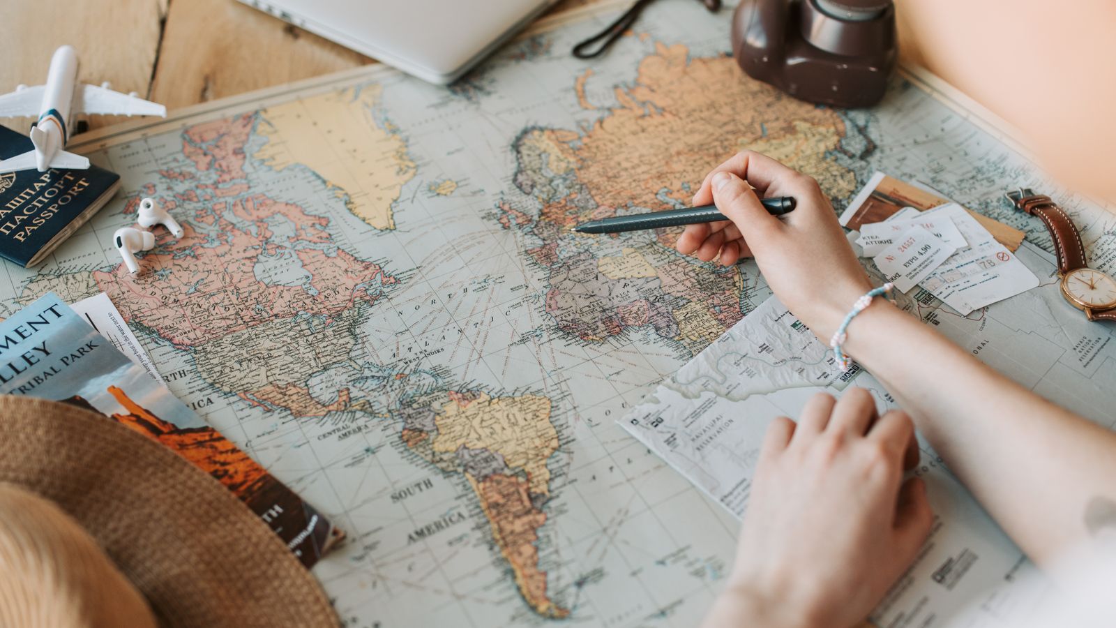 Hands marking destinations on a world map surrounded by a passport, camera and travel essentials