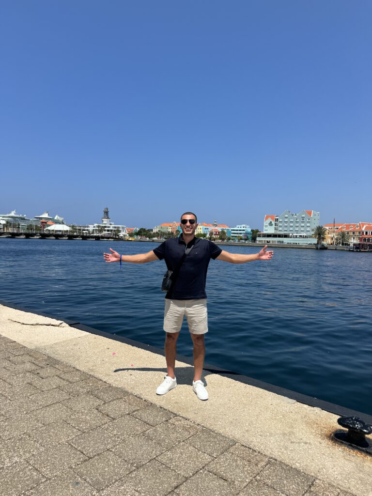 Ryan with arms open on a sunny waterfront lined with colorful buildings